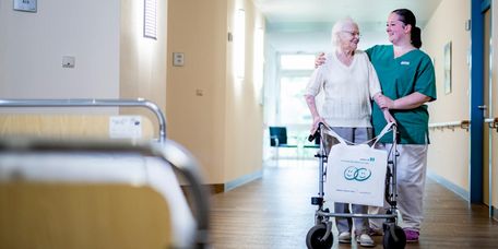 Pflege zum Wohle unserer Patient*innen Pflegekraft begleitet ältere Patientin durch den Flur