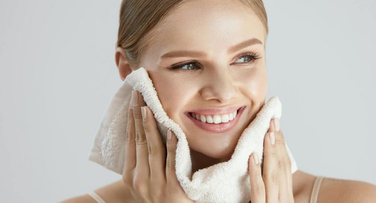 Lächelnde junge Frau reinigt Gesicht mit weißen flauschigen Handtuch