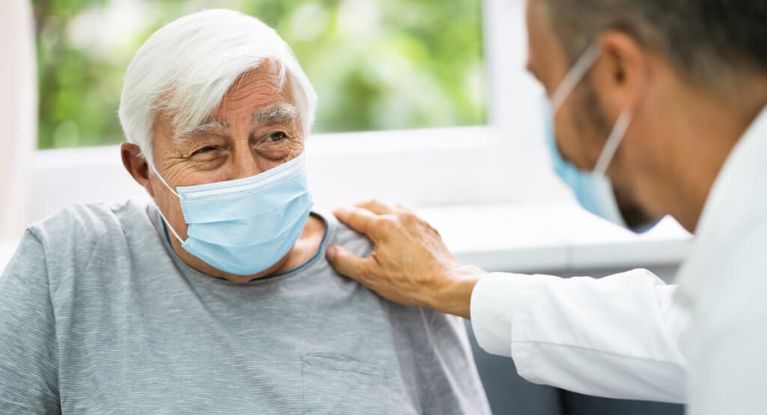 Arzt mit Maske legt einem älteren Patienten mit Maske die Hand auf die Schulter